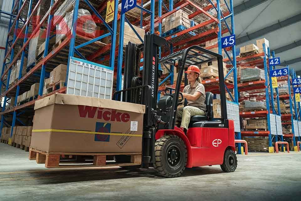 Женщина-оператор управляет вилочным погрузчиком на современном складе, перемещая крупный груз на паллете среди стеллажей с товаром.